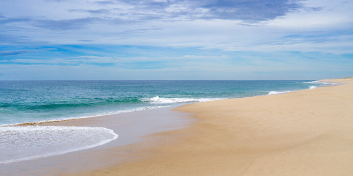 A tranquil sandy beach with gentle waves under a partly cloudy blue sky. Punta Labos Beach, Todos Santo, Mexico