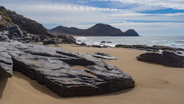 Rocky beach landscape with distant hills under a cloudy sky and waves hitting the shore. Todos Santos, Baja California, Mexico