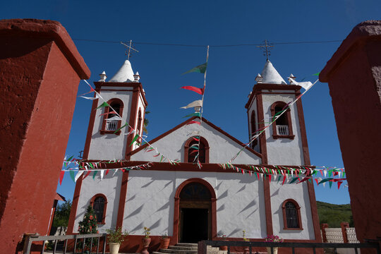 White church with red trim and twin bell towers under a clear blue sky, adorned with festive flags. La Paz, Baja California Sur, Mexico