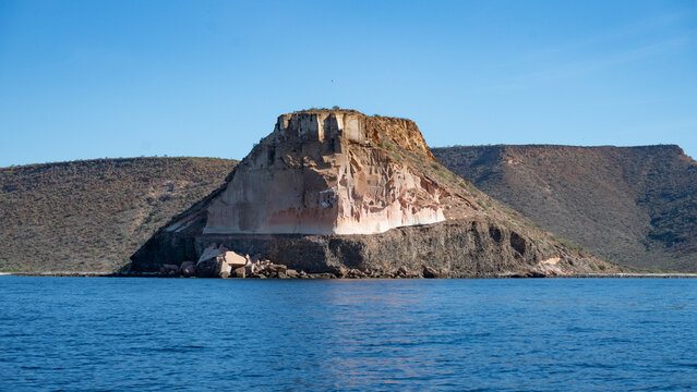 Rocky island rises from the blue ocean under a clear sky with distant desert landscape. Canada