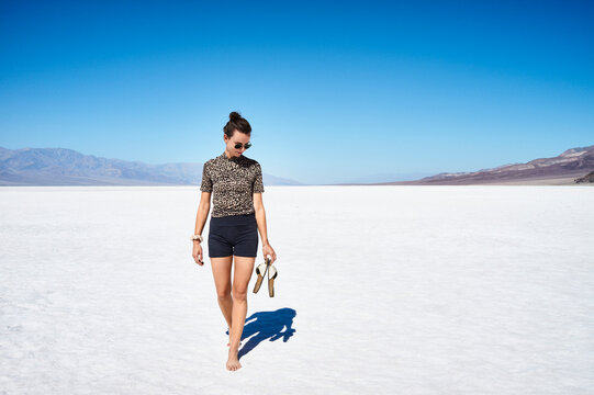 Woman walking barefoot on salt flats under a bright blue sky, holding sandals in hand. Death Valley, California, USA