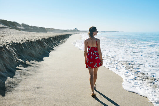 Woman in red floral dress walks along a serene beach with waves gently lapping at shore. Half Moon Bay, California, USA