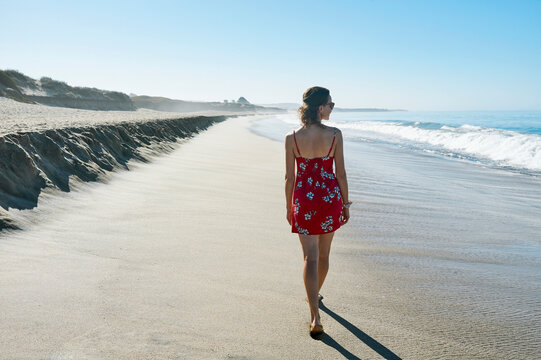 Woman in red dress walks along a serene, sunlit beach with gentle waves and clear skies. Half Moon Bay, California, USA