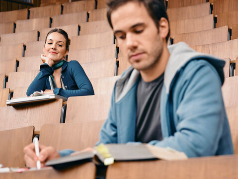Two students in a lecture hall with notebooks, sitting among rows of wooden seats. Germany