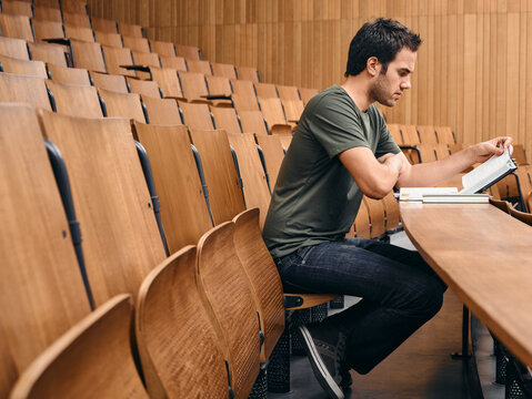 Young man studying in an empty lecture hall with wooden seating and a notepad. Germany
