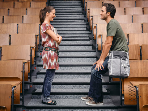 Two adults interact casually on steps in an empty lecture hall with wooden chairs. Germany