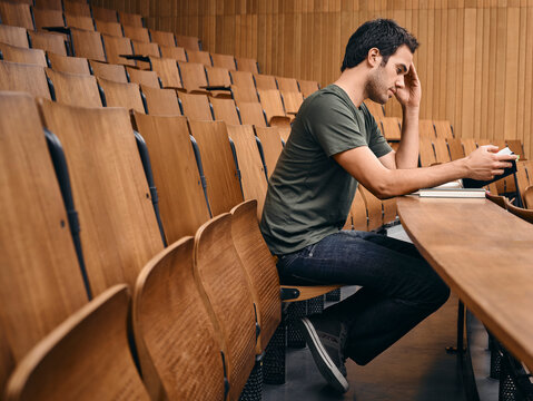 Man sitting in an empty lecture hall using smartphone while reading a book. Germany