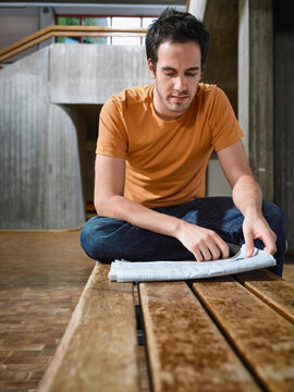 Young man in orange shirt reading a newspaper while sitting on a wooden bench indoors. Germany