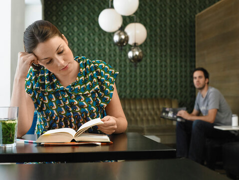 Woman reading a book at a cafe with patterned walls and hanging lights, man looking over to her. Germany