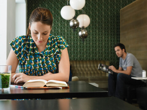 Woman reading a book at a table in a modern cafe with a patterned green wall, man looking over to her. Germany
