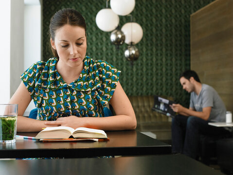 Woman reading a book at a cafe while a man reads a magazine in the background. Germany