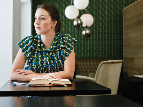 Woman in patterned dress sits at modern table, looking thoughtfully to the side. Germany