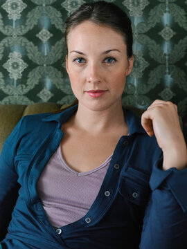 Woman in a blue shirt with a floral wallpaper background sitting on a chair indoors. Germany