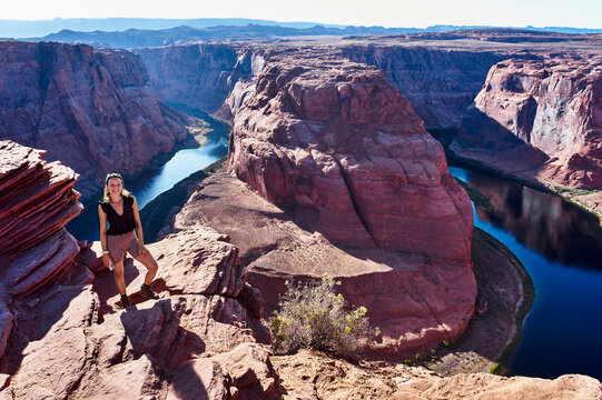 Woman standing on rocky cliff edge overlooking stunning canyon landscape and river below. Horseshoe Bend, Colorado River, Arizona, USA