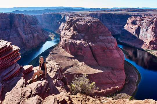 A person sits on a cliff overlooking Horseshoe Bend, with a clear blue sky above the canyon. Horseshoe Bend, Colorado River, Arizona, USA