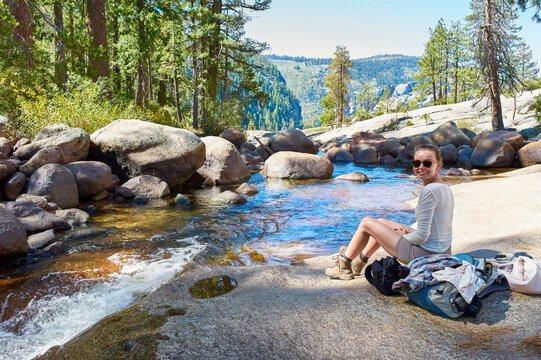Woman sitting by a serene river surrounded by trees and rocks, smiling under a bright sky. Yosemite National Park, California, USA