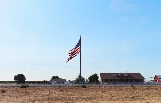 Large American flag waving over rural landscape with house and clear blue sky. California, USA