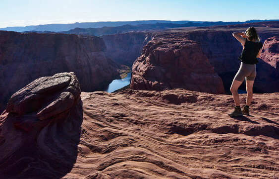Woman standing on a rocky cliff taking a photo of a deep canyon and river below. Horseshoe Bend, Colorado River, Arizona, USA