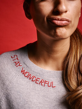 Woman in gray sweater with Stay Wonderful stitching against a red background making a face. Germany