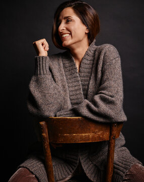 Smiling woman sitting backwards on a wooden chair against a dark background. Germany