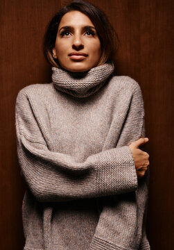 Woman in a cozy turtleneck sweater stands against a wooden background, crossing her arms. Germany