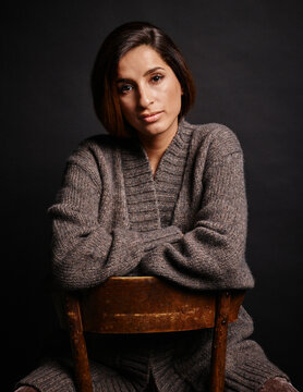 Woman in knitted cashmere cardigan sitting backward on a wooden chair against a dark background. Germany