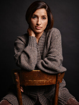 Woman sitting on a wooden chair wearing a cozy knitted cashmere sweater against a dark background. Germany