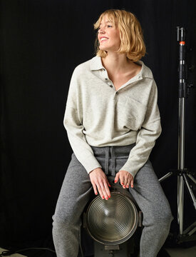 Smiling woman sits on a stage light in casual attire with dark background and studio equipment. Germany