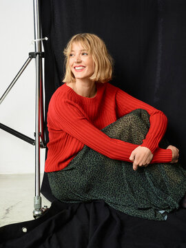 Woman in red sweater and patterned skirt sits smiling against a black backdrop. Germany