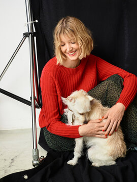 Woman in a red sweater cuddling a small dog on a photoshoot set with a black backdrop. Germany
