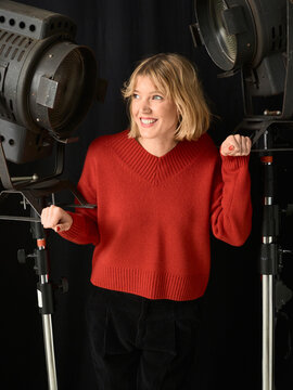 Woman in red sweater smiles between two large studio lights on a black background. Germany