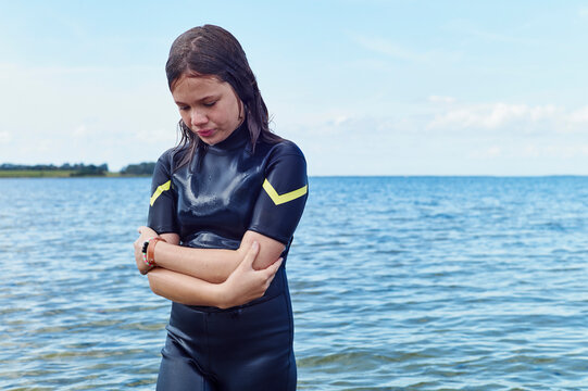 Young girl in wetsuit stands pensively by the calm blue lake on a sunny day. Baltic Sea, Germany