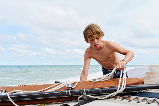 Young boy in swim trunks organizing ropes on a sailboat by the sea under a cloudy sky. Lake Michigan, Illinois, USA
