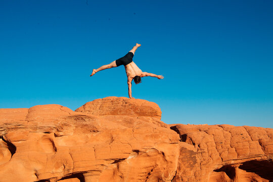 Man performs a one-handed handstand on red rock formations under a clear blue sky. WA, USA