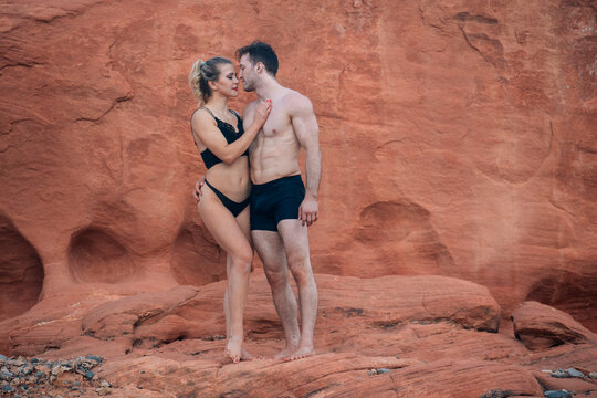 A couple in swimwear embraces against a backdrop of red rocky cliffs. WA, USA