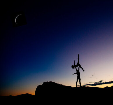 Two silhouettes performing an acrobatic pose on a hill at twilight with a crescent moon. WA, USA