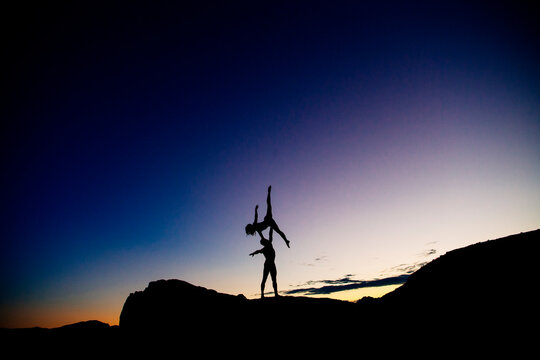 Two people perform an acrobatic pose at sunset on a rocky landscape with a vibrant sky. WA, USA