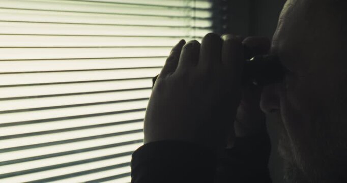 Moody side profile of a man in shadows lifting binoculars to his eyes while hiding behind partially open window blinds. Captures a moment of tension and suspicion.