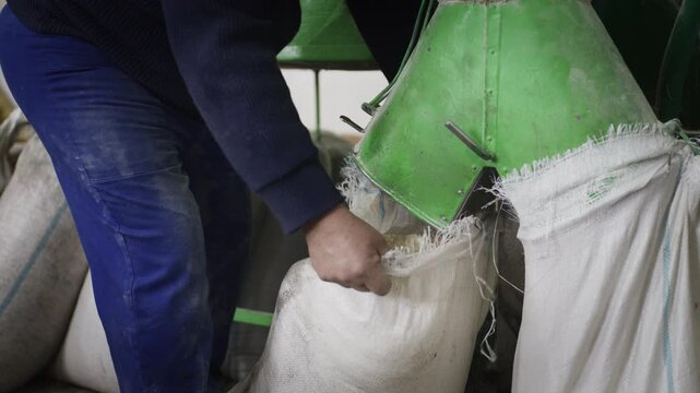 A worker removes a white sack full of processed grain from a green mill agricultural machine