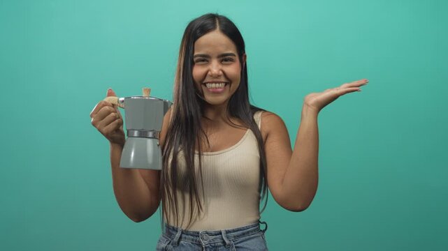 Smiling woman holding espresso maker with open palm presenting in a turquoise studio setting; happiness morning routine.