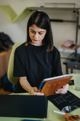 Naklejka premium A woman studies documents while using a tablet and laptop at a tidy home desk. The woman looks focused as she reviews notes in a casual workspace.