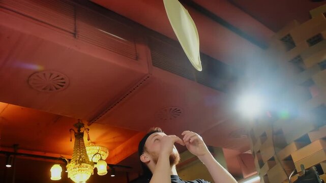 Pizza chef tosses dough disk above head under restaurant ceiling. Dough spins midair while pizza chef tracks flight with raised hands