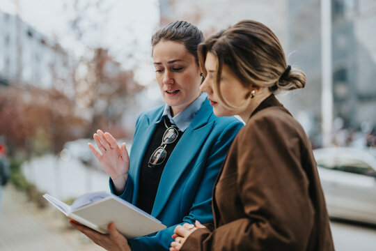 Two women in business attire review a notebook on a city street, engaged in collaboration and discussion.