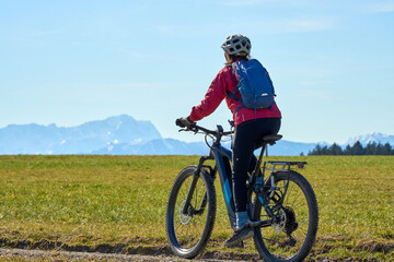 Obraz premium Middle aged woman cycling on an electric bike across green spring meadow in Upper Bavaria with alpine mountain range and Zugspitze silhouette in background under clear blue sky.