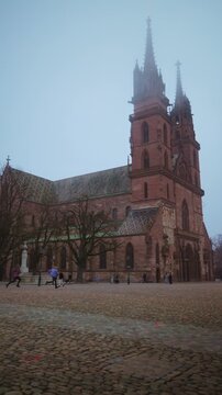 Vertical panning shot of the historic Basel Minster (Basler M&uuml;nster) cathedral under a misty sky, featuring its iconic red sandstone architecture and twin towers.