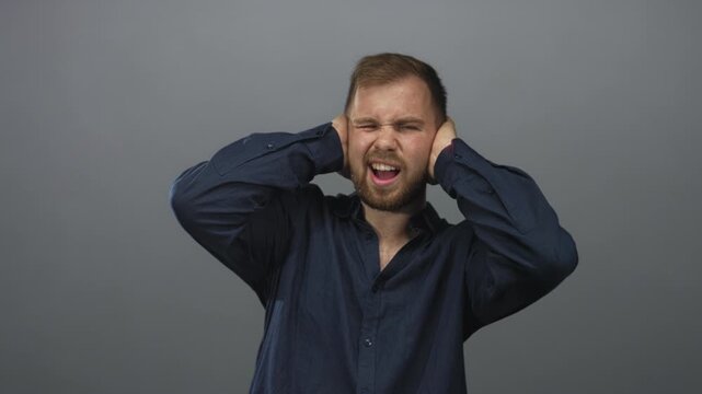 Young caucasian man pressing hands over ears and wincing in gray studio wearing navy shirt; pain noise sensitivity.