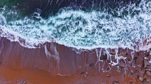 Waves roll onto the sandy beach, creating white foam as they break. The water reflects sunlight. Rocks are visible in some areas as tide rises and falls.