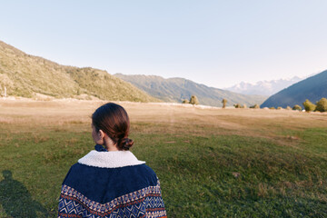 Naklejka premium Woman back portrait in sweater standing in open field, looking at mountain landscape and distant horizon. Nature scene with valley, green grass and clear sky for travel and solitude.