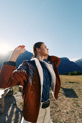 Naklejka premium Woman portrait in mountains outdoors wearing a warm patterned jacket, basking in golden sunlight in an open field. Fashionable traveler enjoying nature, fresh air and mountain landscape.