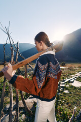 Naklejka premium Woman with guitar outdoors in mountains at sunset, walking along trail wearing patterned sweater and light pants, backlit scene with natural landscape, peaceful travel and music.